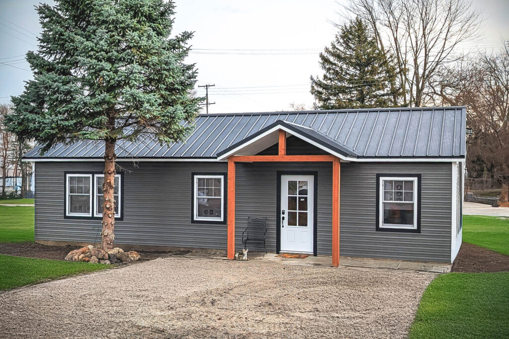 Gray ranch-style tiny home shell with front porch and metal roof, surrounded by grass.