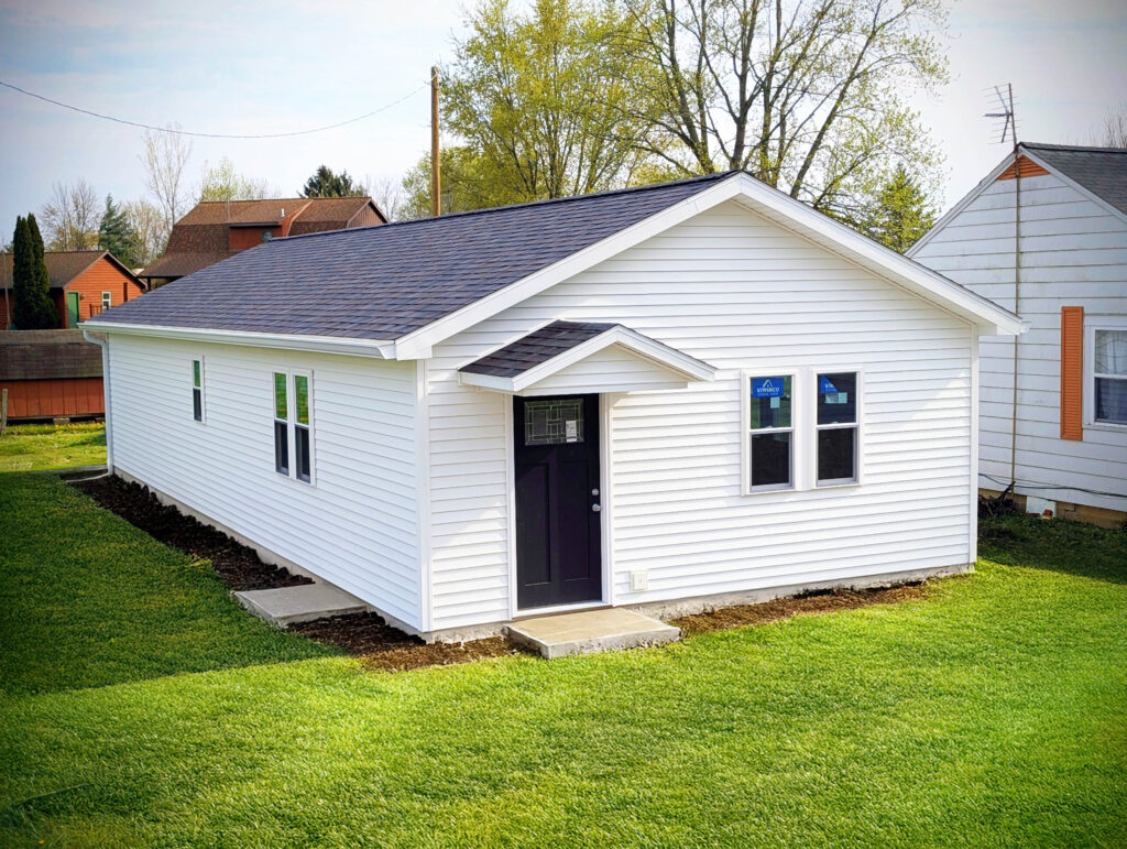 White vinyl-sided tiny home shell with black front door and shingle roof.